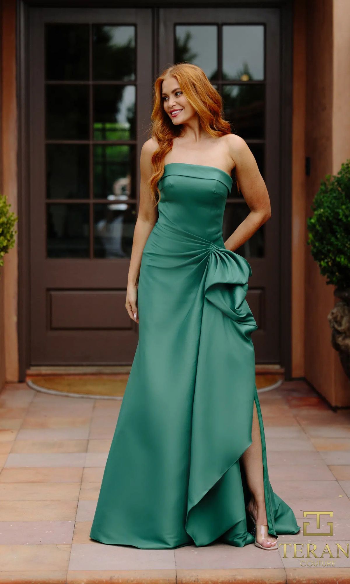 A woman with long red hair stands outside before double doors, wearing a strapless emerald Long Formal Dress: Terani 2111P4019 with a side slit and draped detail.